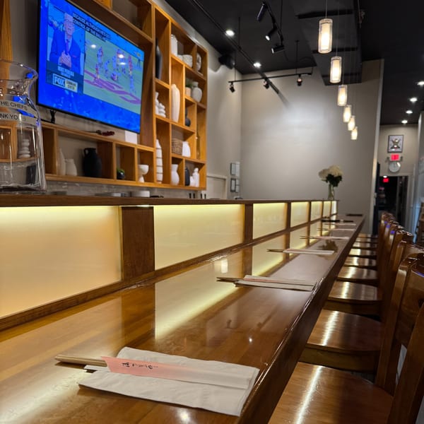 A long, polished wooden sushi bar with chopsticks and chairs, a TV, and decorative shelves.