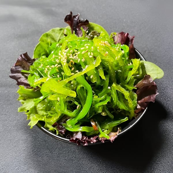 A vibrant green seaweed salad, topped with sesame seeds, served in a black bowl with lettuce.