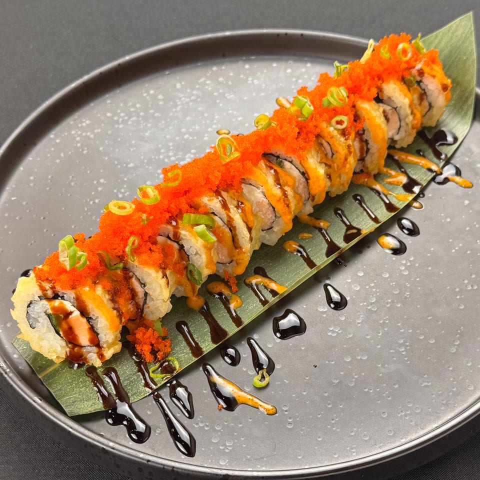 A long sushi roll topped with bright orange roe and green onions, drizzled with sauces on a leaf.
