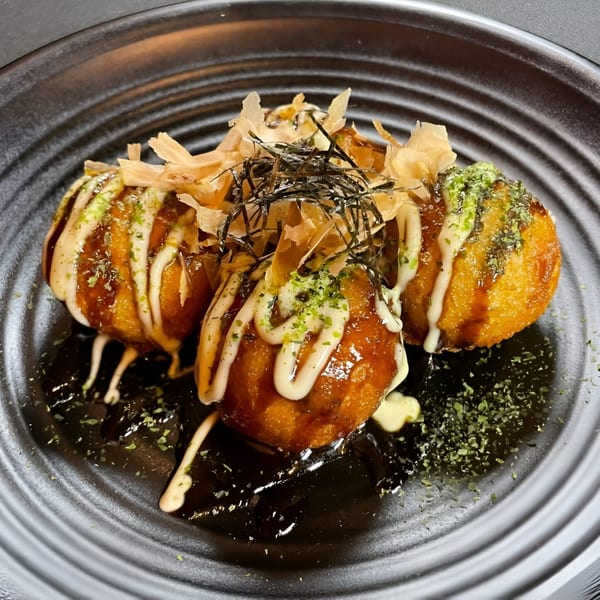 Three golden-brown takoyaki, drizzled with white sauce and topped with bonito flakes and seaweed, on a dark plate.